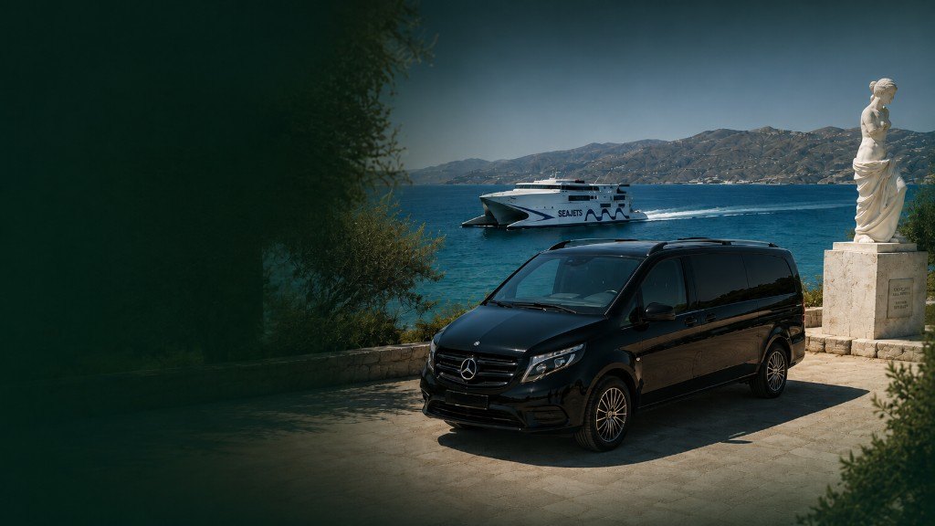 Black Mercedes-Benz V-Class on a terrace in Milos, with the sea, a ferry and the island coastline beyond. Vehicle registration is obscured.